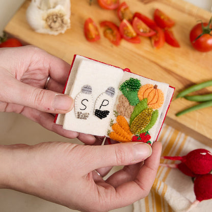 Miniature felt ornament shaped like a cookbook, hand-felted and embroidered with cooking-themed details including salt, pepper, vegetables, and a stovetop. Crafted from merino-blend wool with soft pages and a hanging string, this ornament measures approximately 2.5 inches tall. A cozy tribute to home cooking and culinary tradition.  Open to pages with salt and pepper and vegetable.