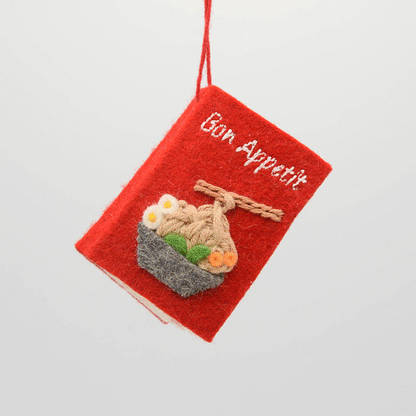 Miniature felt ornament shaped like a cookbook, hand-felted and embroidered with cooking-themed details including salt, pepper, vegetables, and a stovetop. Crafted from merino-blend wool with soft pages and a hanging string, this ornament measures approximately 2.5 inches tall. A cozy tribute to home cooking and culinary tradition. GIF 360 degree book