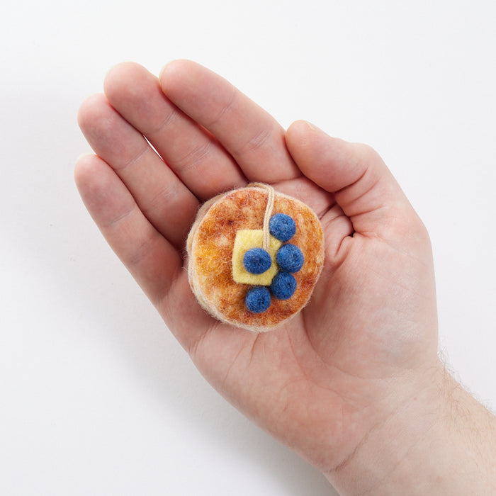 Craftspring’s Blueberry Pancake Stack Ornament from the Iconic Craftspring Ornaments Bundle displayed in front of hand to show its size — felt stack of pancakes with embroidered and beaded details, a charming heirloom-quality piece for Christmas gifting — ethically crafted by artisans using sustainable wool.