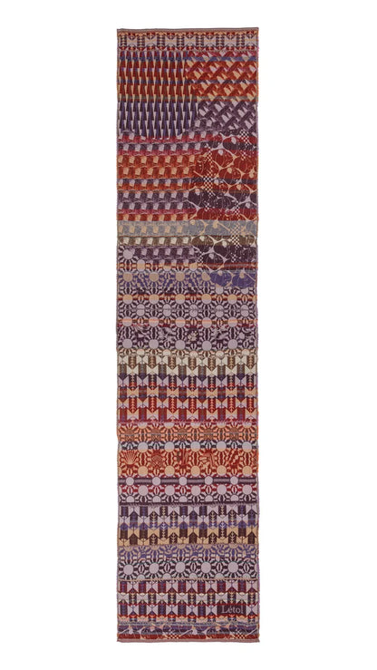 Létol Come Scarf in Cassis, laid flat and fully extended against a white background, showing the full length of the scarf with a rich gradient composition — the top third featuring bold diagonal chevron and lattice bands in russet, burgundy, and terracotta, transitioning through a central zone of repeating geometric rosette and medallion motifs in violet and mauve on a dusty rose ground, deepening into alternating bands of blackcurrant chevrons, floral rosettes, and Art Deco fan patterns with the woven Léto