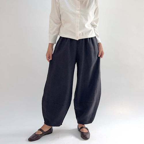Model wearing the Bryn Walker Heavy Linen Oliver Pant in Fig, angled front view showing barrel-leg silhouette, elastic waistband, side pocket, and tapered ankle in deep fig heavy linen, styled with cream button-up and brown Mary Jane flats