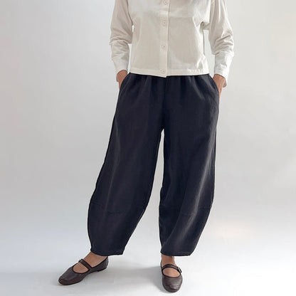 Model with hands in pockets wearing the Bryn Walker Heavy Linen Oliver Pant in Fig, full front view showing barrel-leg silhouette, elastic waistband, side pockets, tapered ankle, and deep fig heavy linen, styled with cream button-up and brown Mary Jane flats