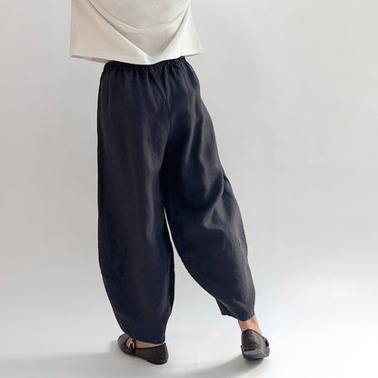 Back view of model wearing the Bryn Walker Heavy Linen Oliver Pant in Fig, showing elastic waistband, full barrel-leg drape, tapered ankle, and deep fig heavy linen texture, styled with cream top and brown Mary Jane flats