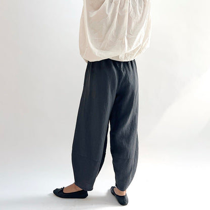 Back view of model wearing the Bryn Walker Heavy Linen Oliver Pant in Moonlight, showing elastic waistband, full barrel-leg drape, and tapered ankle in deep moonlight charcoal heavy linen, styled with cream oversized top and black ballet flats