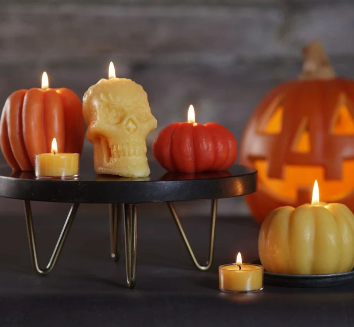Big Dipper Wax Works Halloween candle collection displayed on a black metal hairpin-leg stand, showing two dark orange ribbed beeswax pumpkin candles and a natural beeswax skull candle with lit beeswax tealights, alongside a large natural beeswax pumpkin candle on a black dish in the foreground, with a glowing carved jack-o-lantern in the background