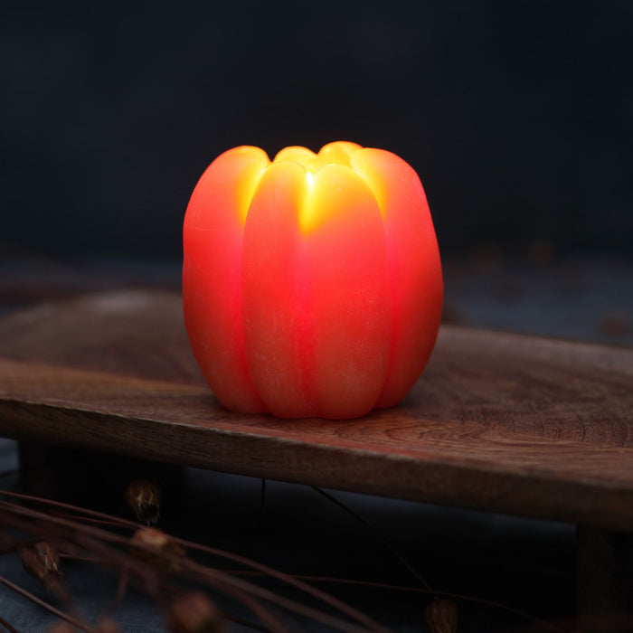 Big Dipper Wax Works Medium Beeswax Pumpkin Candle in light orange, lit and glowing on a rustic wooden surface, showing a tall upright ribbed pumpkin shape with warm amber light glowing through the wax from an internal flame, against a dark black background