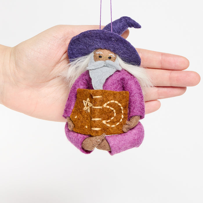 Hand-felted wool ornament of a grey-bearded wizard wearing a pointed hat, holding an open spell-book beneath swirling stardust. Featuring a warm-toned complexion, white hair, and flowing robes, this whimsical figure is crafted from natural merino-blend wool. Includes a hanging string and measures approximately 5 inches tall.