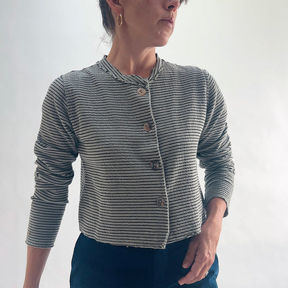 Close front view of model wearing a squirrel and black fine horizontal stripe cropped cardigan, showing the asymmetrical button closure, band neckline, and crinkle-texture stripe detail, styled with dark teal wide-leg pants.