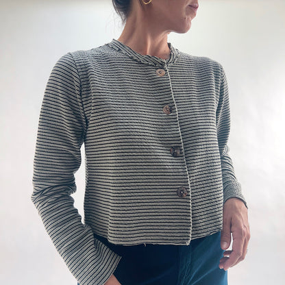 Three-quarter front view of model wearing a squirrel and black fine horizontal stripe cropped cardigan with asymmetrical button closure and long sleeves, looking to the side, showing the relaxed cropped silhouette and crinkle-texture fabric, styled with dark teal wide-leg pants.